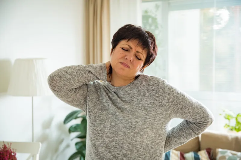 mature woman suffering from backache at home massaging neck with hand, feeling exhausted, standing in living room