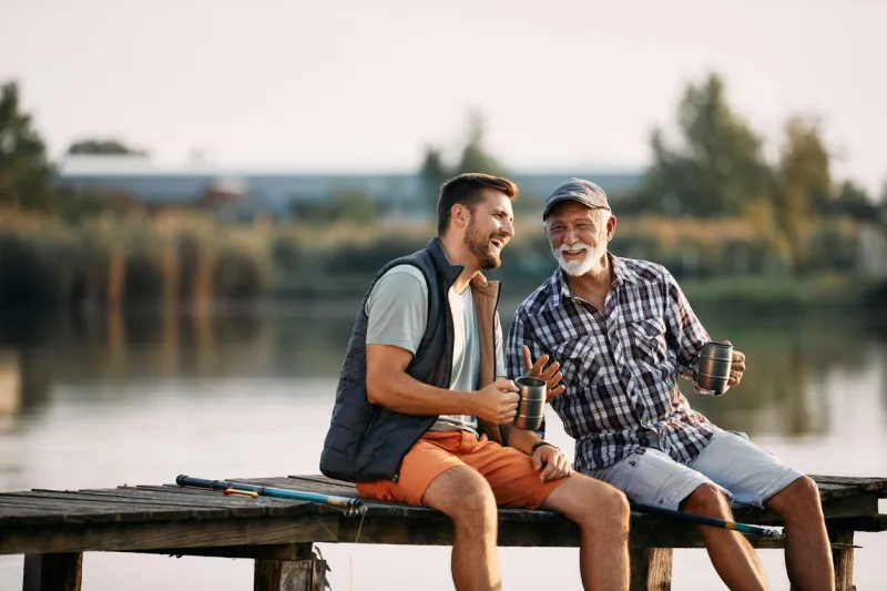 happy senior fisherman and his adult son talking while having a drink on a pier copy space