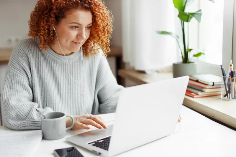 portrait of curly haired redhead caucasian female in wired headphones spending leisure time using laptop during coffee break, sitting at kitchen table with smartphone, visiting popular web pages