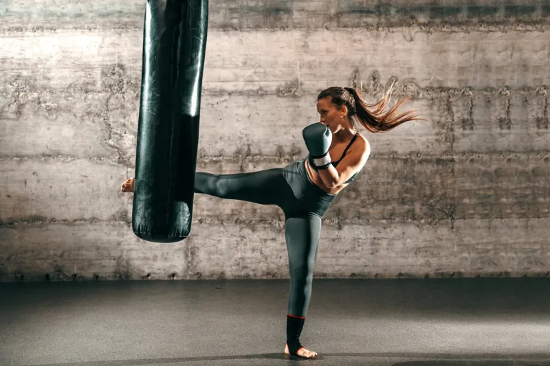 dedicated strong brunette with ponytail, in sportswear, bare foot and with boxing gloves kicking sack in gym
