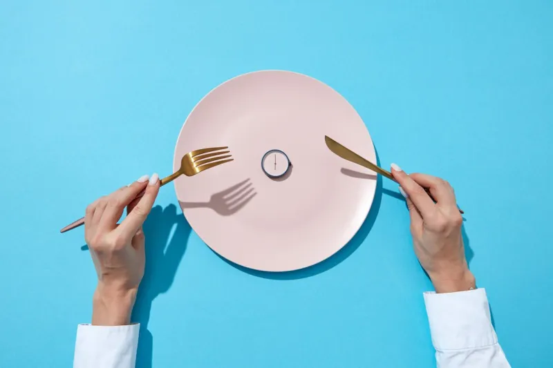 round watch of six o'clock and woman's hand with fork and knite in agirl's hands on a blue background with shadows time to lose weight, eating control or diet concept place for text