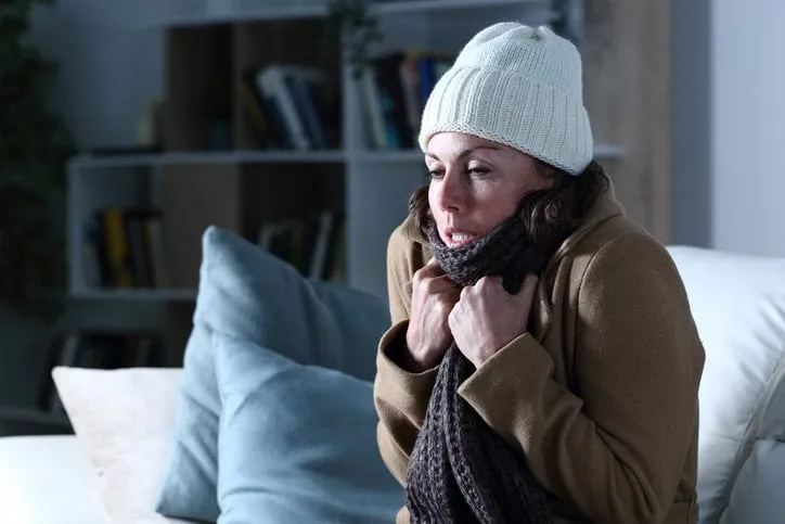 cold adult woman covered with clothes freezing sitting on the sofa at night at home