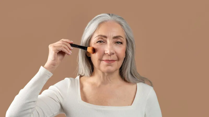 natural makeup for mature skin charming senior lady putting on organic cosmetics with brush on brown studio background, banner design elegant older woman applying blusher or powder