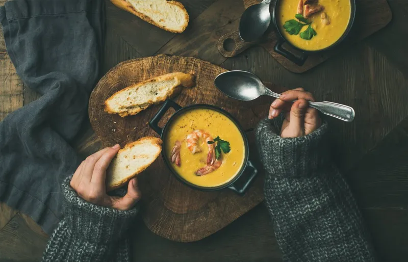 woman in warm grey sweater eating corn creamy chowder soup with prawns served in individual pots, top view woman' s hand keeping spoon and bread slice flat-lay of rustic dinner table slow food, winter warming food concept