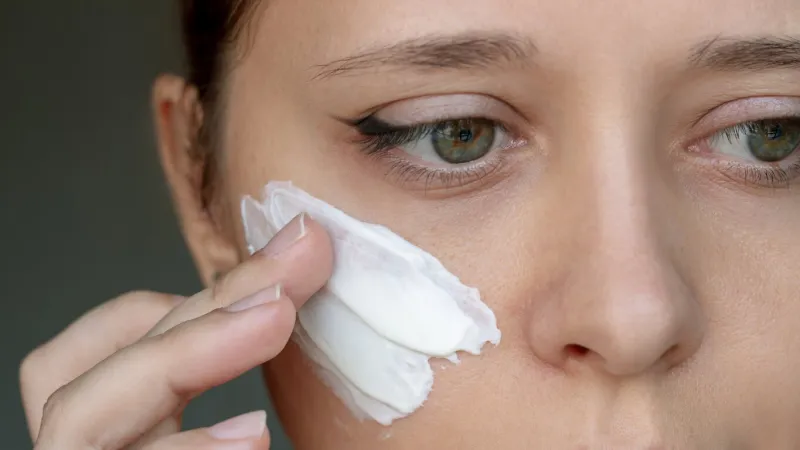 close up of a young caucasian woman applying a thick smear of cream touching the face with her hand on a dark background nourishing moisturizing cream cosmetology and beauty, skin care