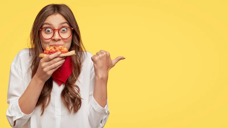 lunch time concept attractive cheerful woman eats delicious italian pizza, surprised with wonderful taste, indicates with thumb aside, shows where pizzeria situated, suggests to visit and have snack