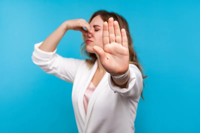 bad smell portrait of woman with wavy hair in white jacket pinching her nose and showing stop gesture, expressing disgust to unpleasant odor, fart gases, her eyes closed with abhor blue background