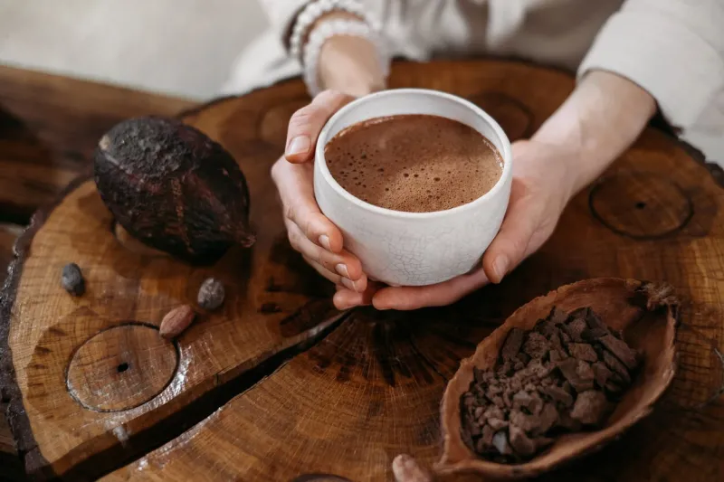 hot handmade ceremonial cacao in white cup woman hands holding craft cocoa, top view on wooden table organic healthy chocolate drink prepared from beans, no sugar giving cup on ceremony, cozy cafe