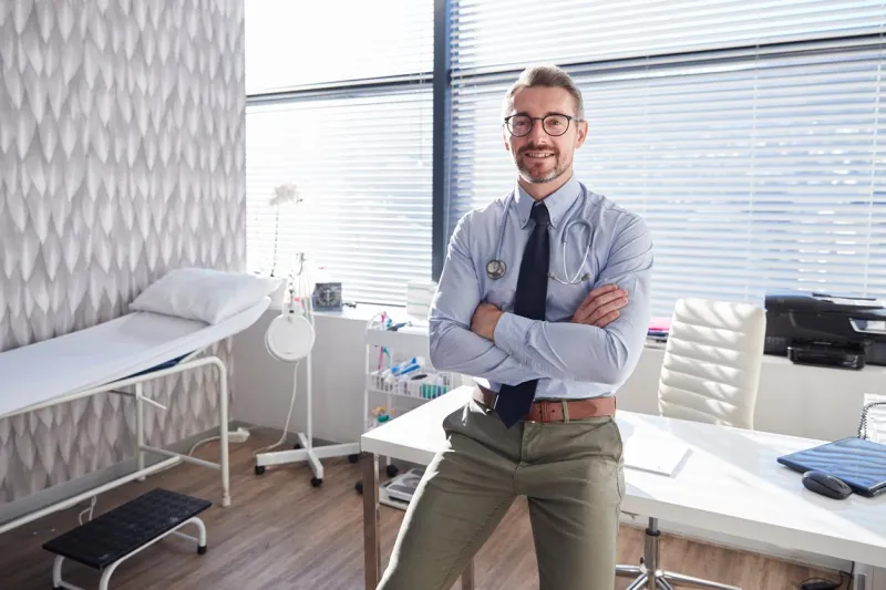portrait of smiling mature male doctor with stethoscope standing by desk in office