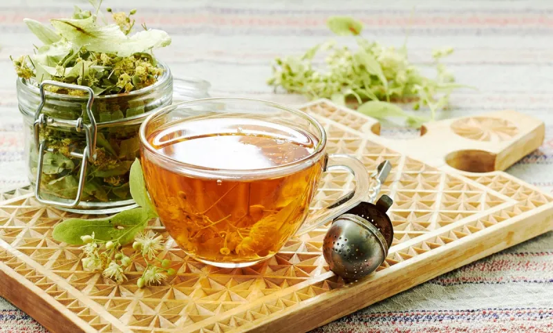 linden cup of herbal healing tea with lime tree bloom and glass can with dried herb nearby on rustic wooden cutting board, backlit, closeup, copy space, alternative medicine and naturopathy concept