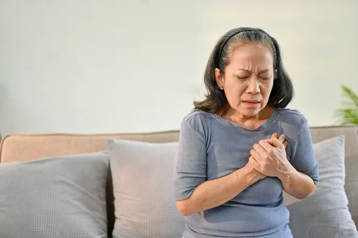 unwell and sick elderly asian woman sits on sofa in her living room, touching her chest, suffering from heart attack or chest pain health problems concept
