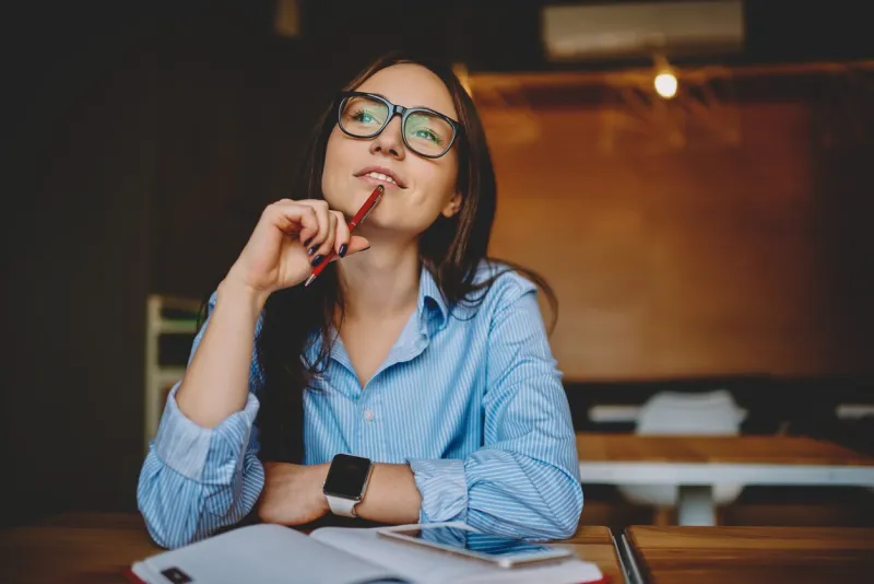 dreamy woman podring while working on journalistic publication sitting with notebook in cafe,thoughtful female student in eyewear doing homework task solving problems and analyzing information