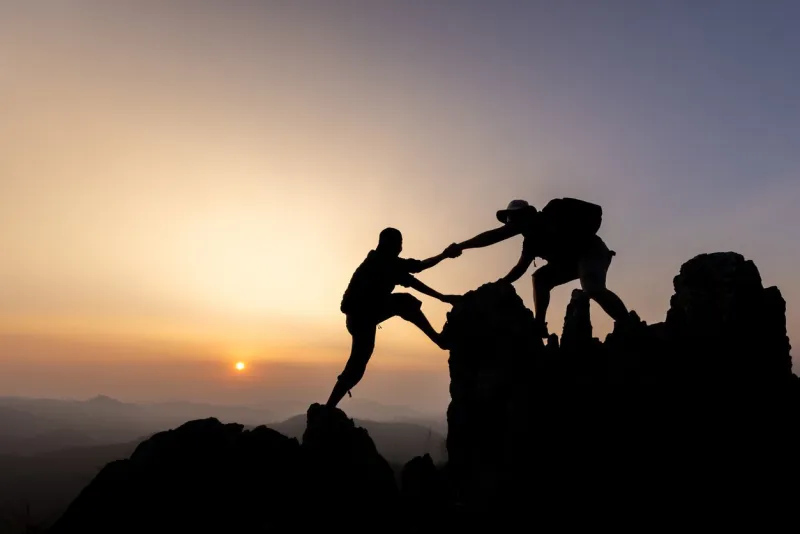 silhouette of person hikers climbing up mountain cliff and one of them giving helping hand people helping and, team work concept