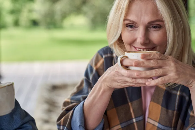 smiling attractive mature female is drinking tea in open terrace of house among green nature