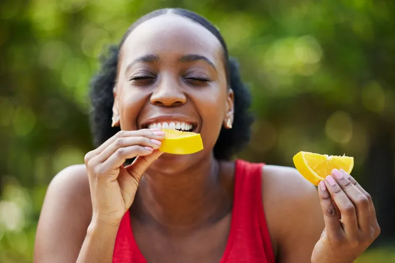 black woman, vitamin c and eating orange slice for natural nutrition or citrus diet in nature outdoors happy african female person enjoying bite of organic fruit for health and wellness in the park