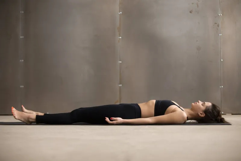 young attractive woman practicing yoga, lying in dead body, savasana exercise, corpse pose, working out, wearing black sportswear, cool urban style, full length, grey studio background