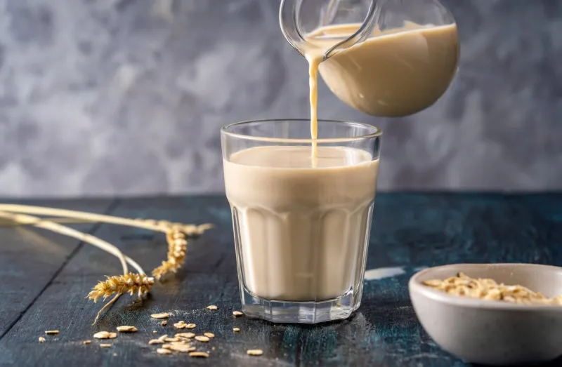 oat milk being poured from a jug into a transparent glass on a wooden surface, with oat flakes and wheat stalks around