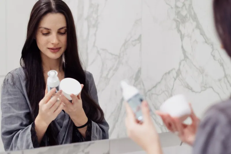 attractive young woman with perfect long hair looking at bottle of face cleansing gel and jar with moisturizing cream during her morning routine at bathroom wellness and selfcare concept