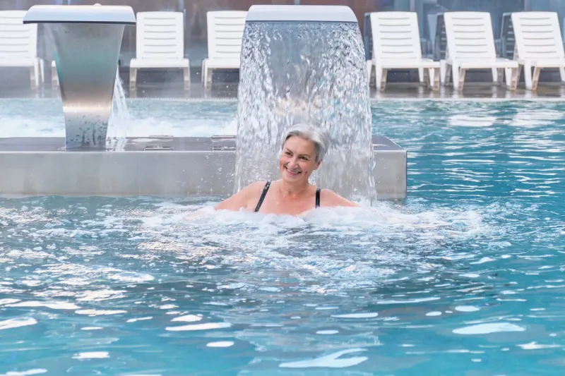 senior caucasian smiling woman with gray hair enjoying falling jets of water hydromassage in outdoor thermal swimming pool mental health concept