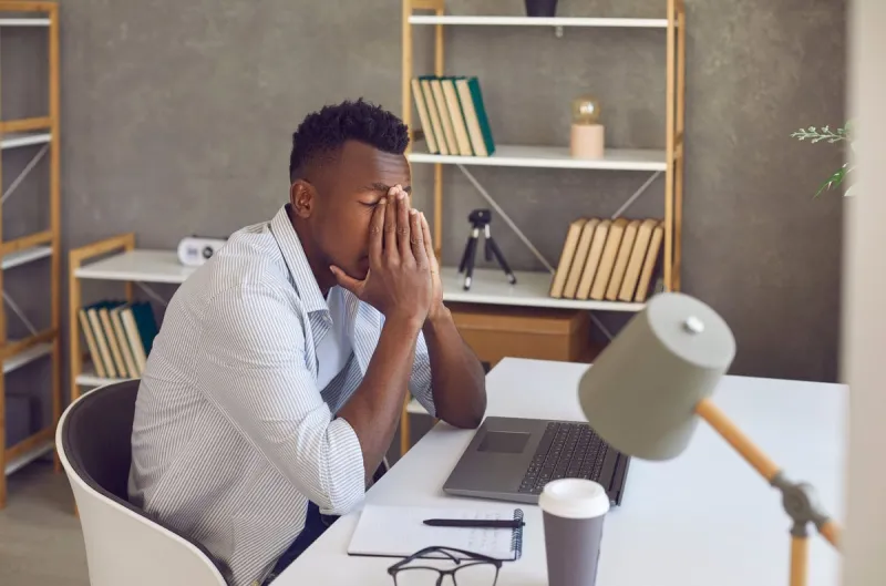upset black man overwhelmed with problems, financial difficulties or stress and negativity at work tired and frustrated male student or office worker covers face sitting at desk with laptop computer