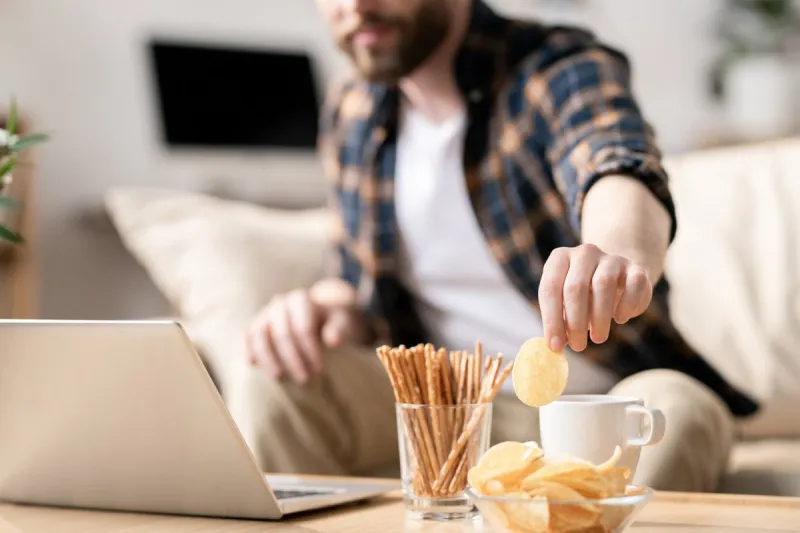 young man taking potato chip out of glass bowl while sitting on sofa in front of laptop on table and having snack