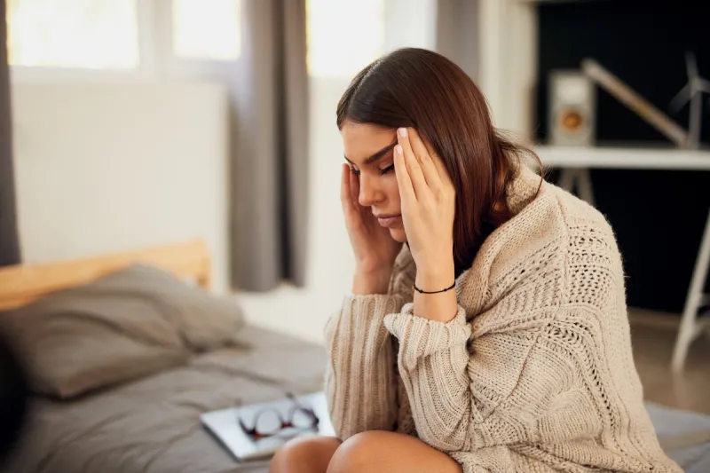attractive caucasian brunette dressed in beige sweater sitting on bed in bedroom and having headache