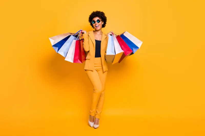 full length photo of positive bruntte hair dark skin girl feel enthusiastic, hold bags addicted shopper shopping on leisure time wear style outfit sunglass high-heels isolated yellow color background