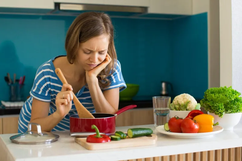 closeup portrait of dissatisfied young beautiful woman looking into sauce pan and cooking in kitchen tasteless dish concept front view