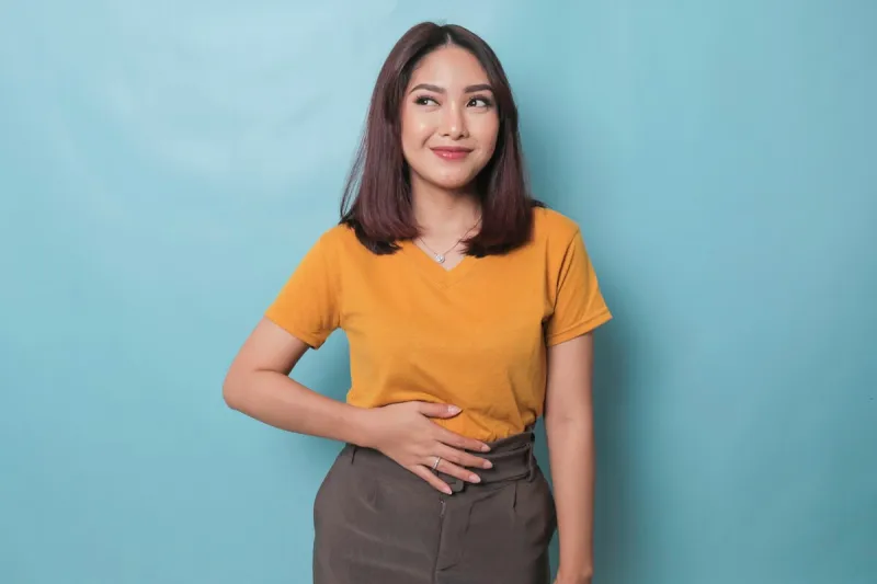 pleased cheerful asian woman keeps hand on belly feels full after delicious dinner dressed casually stands thoughtful against blue background