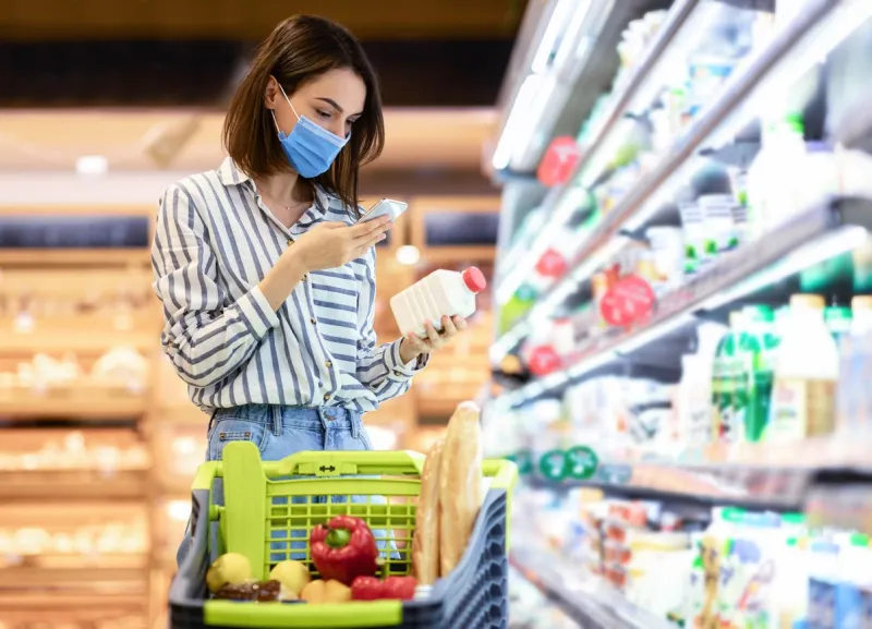 young woman in disposable face mask taking dairy products from shelf in the supermarket, holding bottle and smartphone, scanning bar code on product through mobile phone, walking with trolley cart