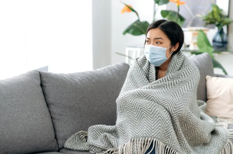 the concept of illness and self-isolation during influenza or covid19 young sick tired asian woman sits upset on sofa at home, wearing medical protective mask, covered with blanket, looks away sadly