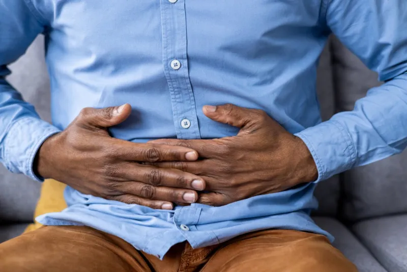 close-up of man's hands clutching stomach indicating abdominal pain and discomfort concept of digestive issues, stomach ache, and health problems man in blue shirt and brown pants sitting on couch