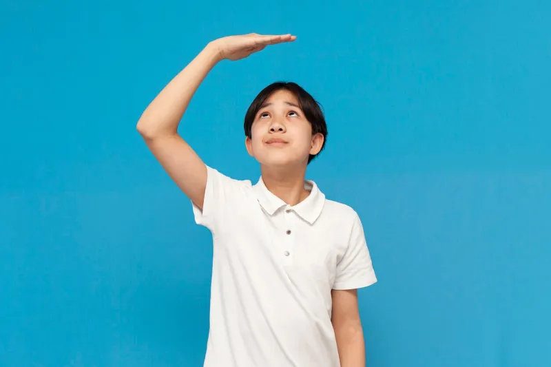 asian boy of twelve years old measures his height with his hand on blue isolated background, korean not tall child dreams of growing up and holds his hand above his head