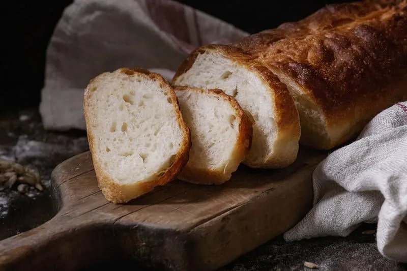 tranche de pain de blé blanc fait maison avec graines de blé et farine sur planche à découper en bois et serviette en lin blanc sur table de cuisine en bois foncé de style rustique