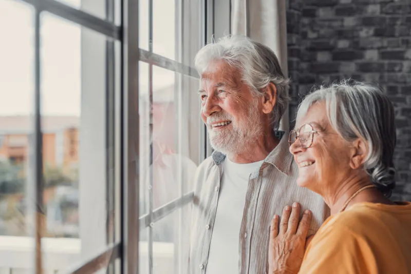 happy bonding loving middle aged senior retired couple standing near window, looking in distance, recollecting good memories or planning common future, enjoying peaceful moment together at home