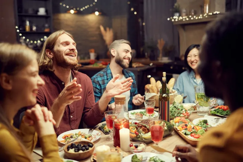group of emotional young people enjoying dinner party with friends and smiling happily sitting at table in dimly lit room, copy space
