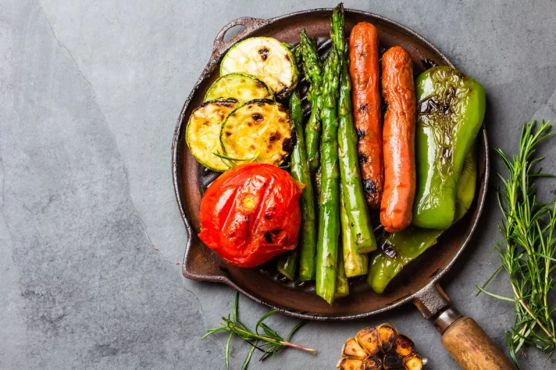 grilled green vegetables - zucchini, asparagus, bell pepper, garlic, sausages, lemon and rosemary on cast iron grill pan gray slate background top view