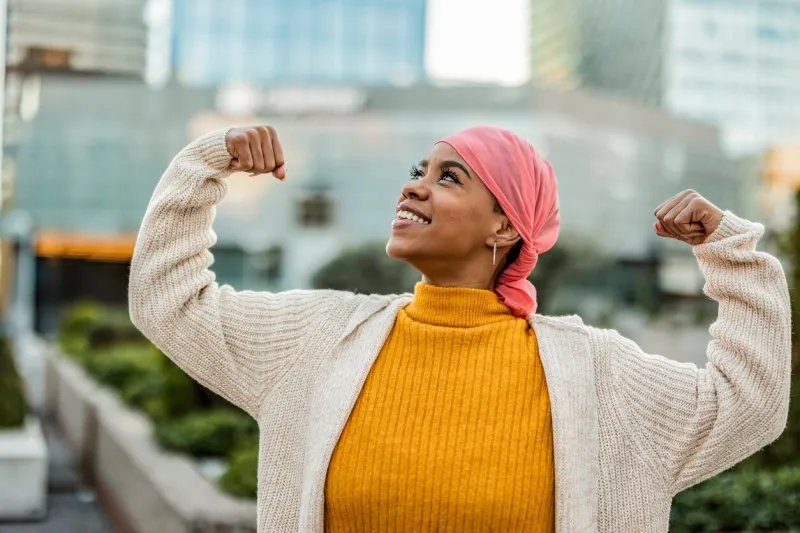 joyful cancer survivor in her 30s, wearing a pink headscarf and yellow sweater, flexing her muscles in an urban setting