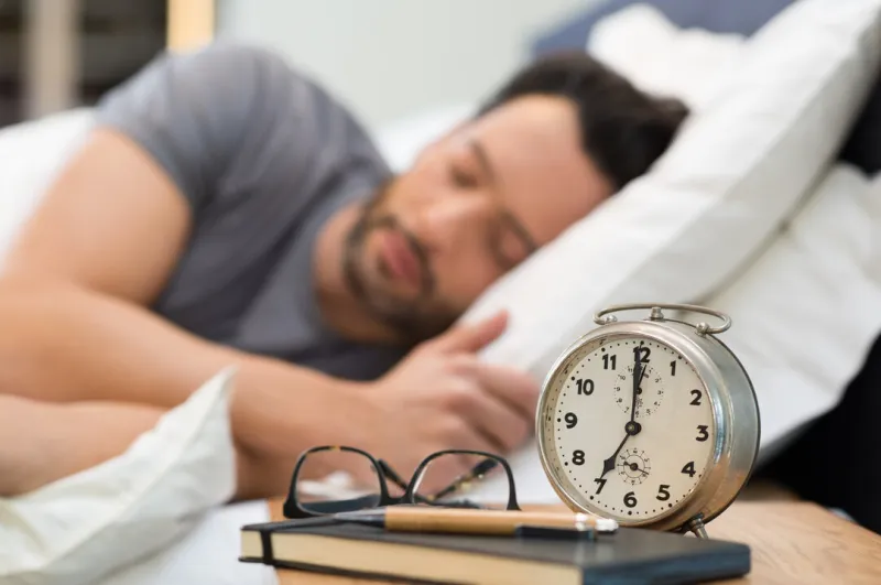 young man sleeping in his bedroom man sleeping with an alarm clock in foreground a calm man in his bed before waking up in his room close up of alarm clock on bedside table
