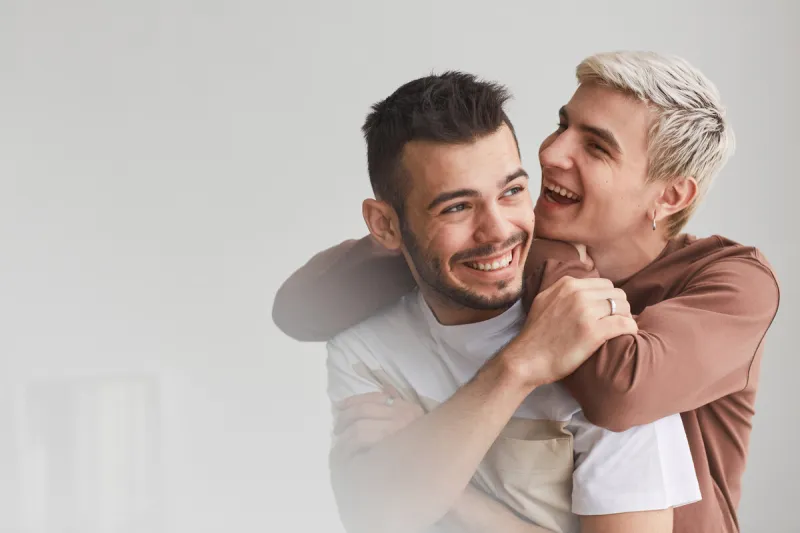 candid waist up portrait of carefree gay couple embracing indoors and laughing happily while posing against white, copy space