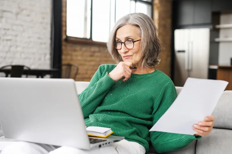 serious senior accountant attentively looking at laptop screen, examines the budget, documents of the company, on the living room, holding laptop on lap, working online, prepares an annual report