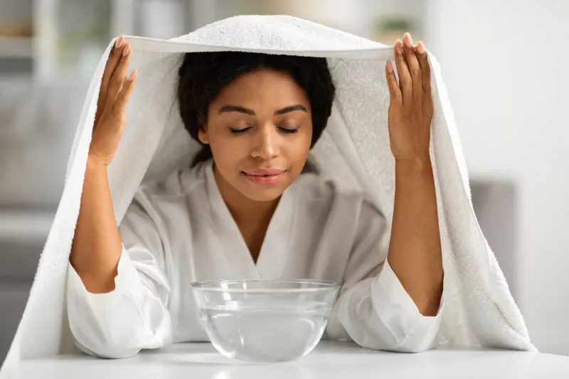 beautiful black woman covered with towel inhaling hot steam at home, attractive young african american female doing inhalations over bowl while making beauty skincare routine, closeup shot