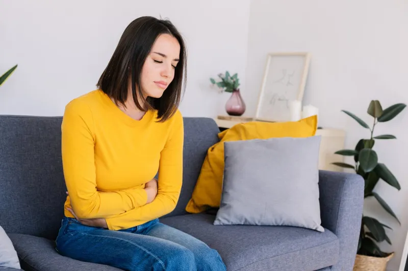young woman suffering stomach ache or abdominal pain while sitting on couch at home menstruation or pregnancy health care effects