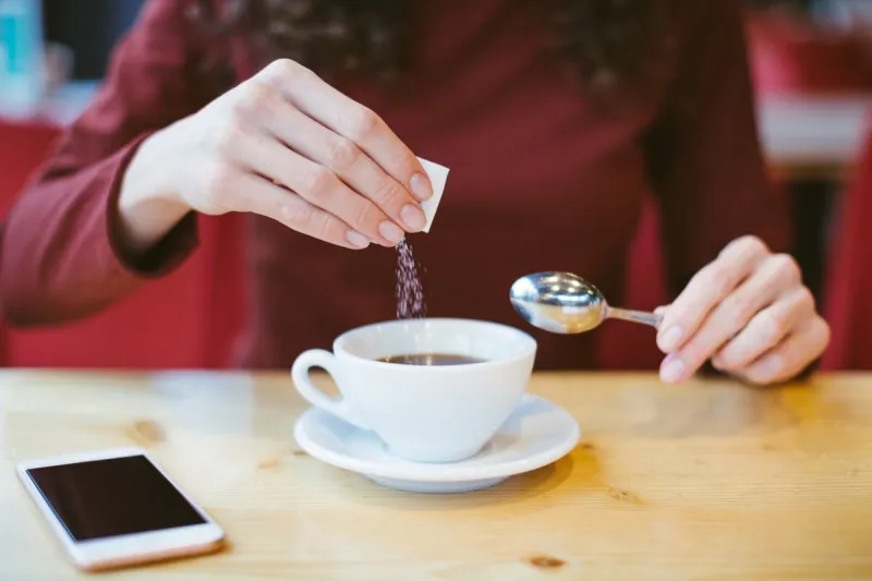 woman's hands pouring sugar into black coffee - girl sitting at the table with espresso and smartphone - blood and glycemic index control for diabetes -excess of white sugar in food concept