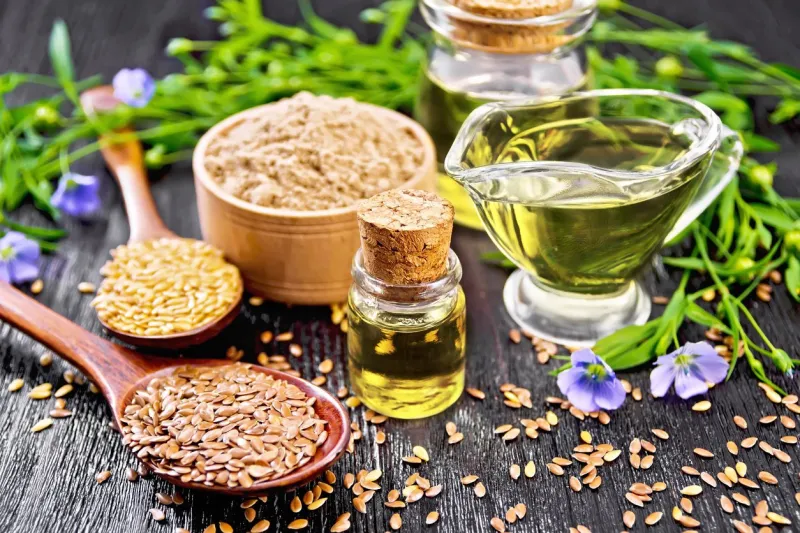 linseed oil in two glass jars and a gravy boat with white and brown flax seeds in spoons, flour in a bowl, leaves and flowers on black wooden board background