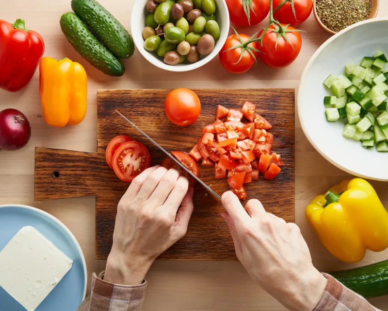 step by step recipe for salad horiatiki, wooden board for cutting vegetables and ingredients hands with knife, chopping tomatoes
