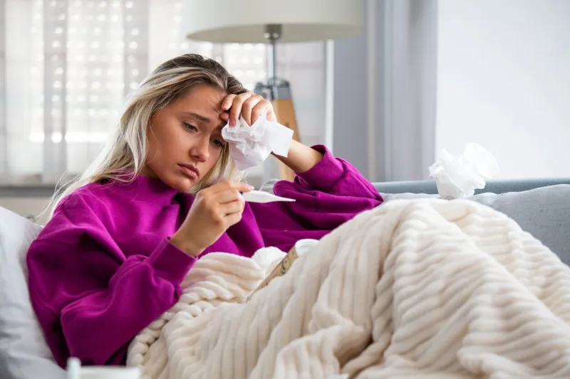 sickness, seasonal virus problem concept woman being sick having flu lying on sofa looking at temperature on thermometer sick woman lying in bed with high fever cold flu and migraine