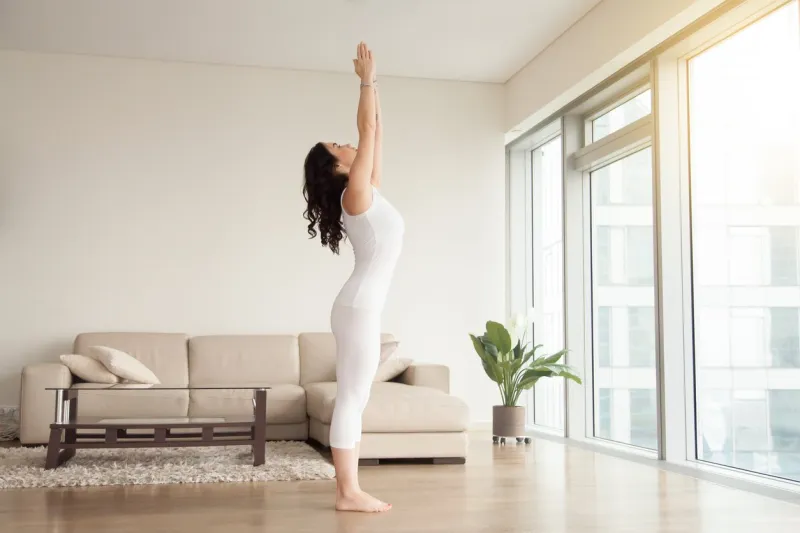 young attractive woman practicing yoga at home, standing in mountain exercise, tadasana pose, working out, wearing white clothes, indoor full length, in the center of living room