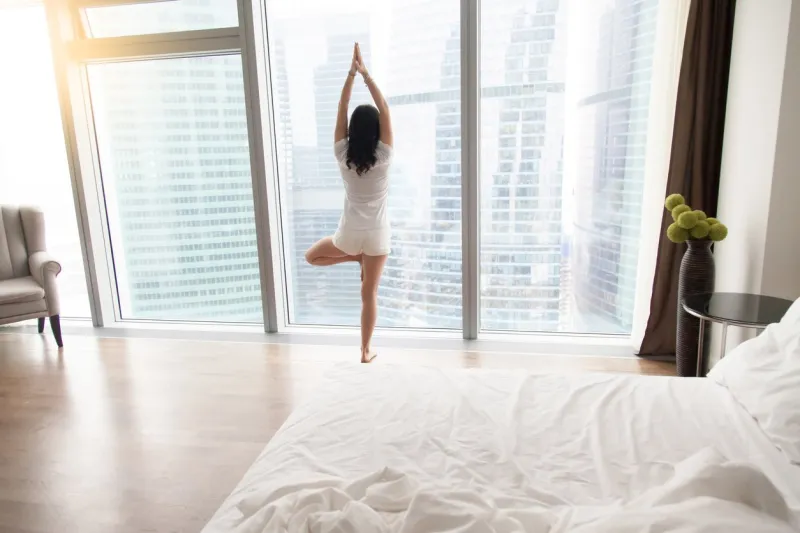 young attractive woman practicing yoga at home after waking up, standing in vrksasana exercise, tree pose, working out, wearing white casual clothes, indoor full length, bedroom, unmade bed, rear view