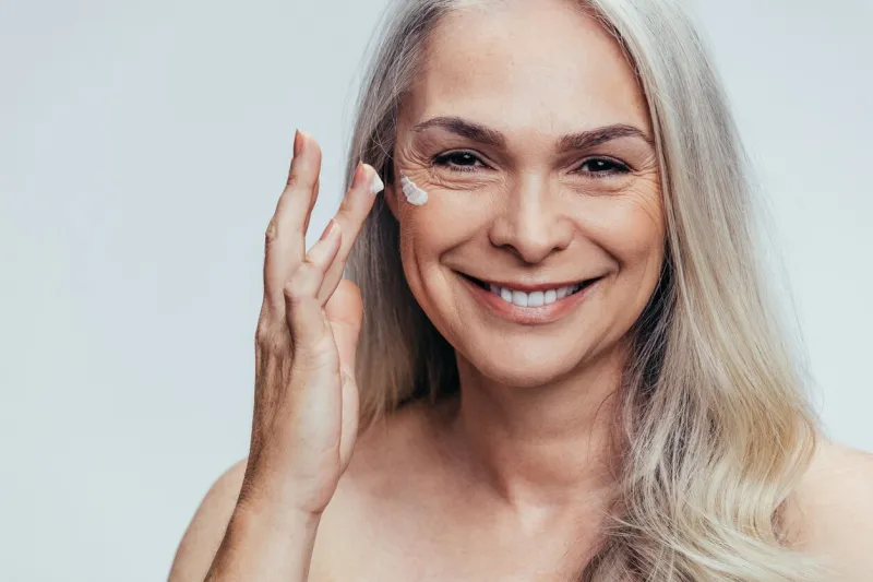 smiling mid adult caucasian woman applying anti aging cream on her face senior female woman applying moisturizer on her face against grey background